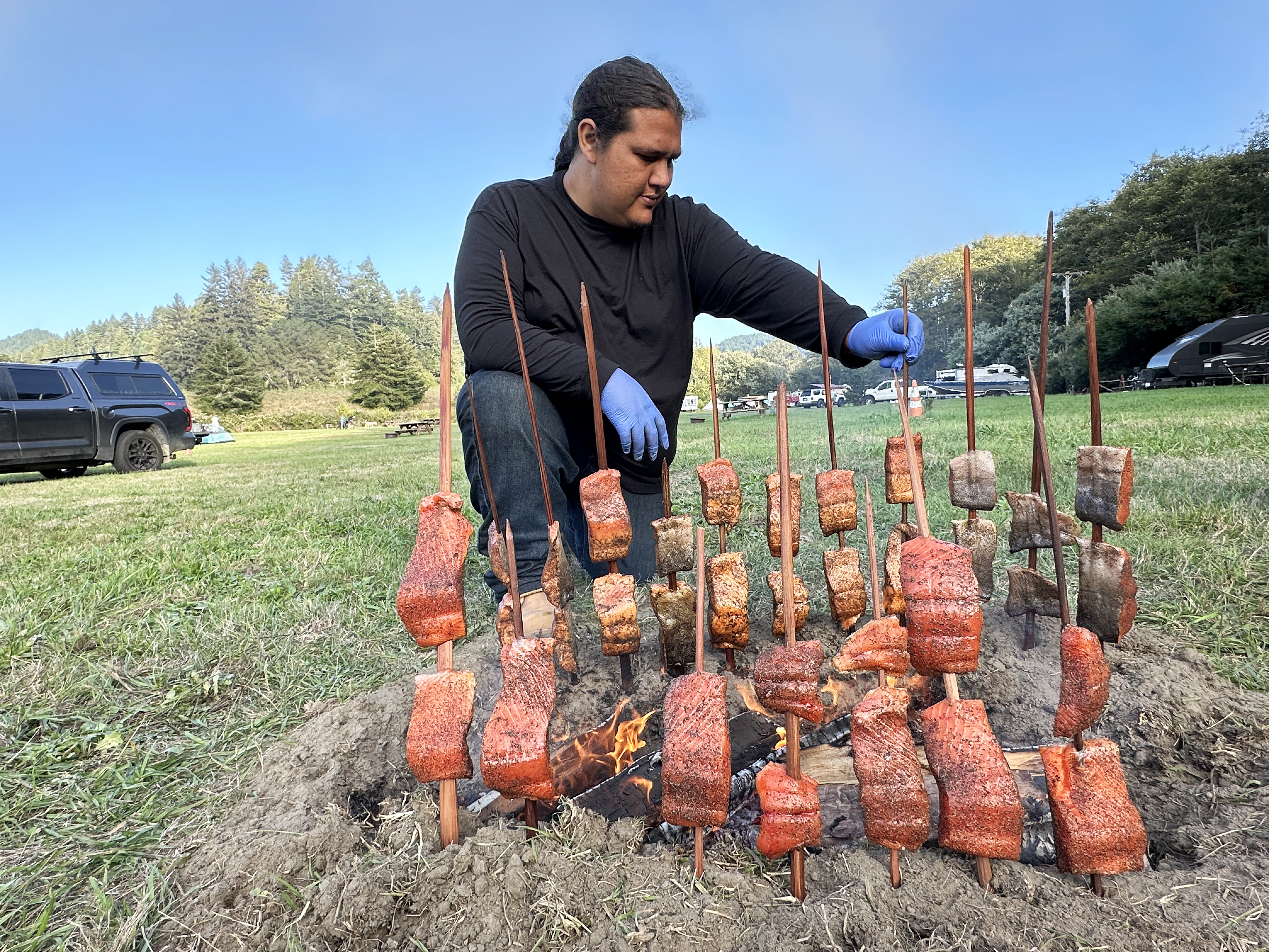 Man crouches next to several pieces of salmon skewered on sticks; the sticks are stuck into the ground outdoors around a fire.