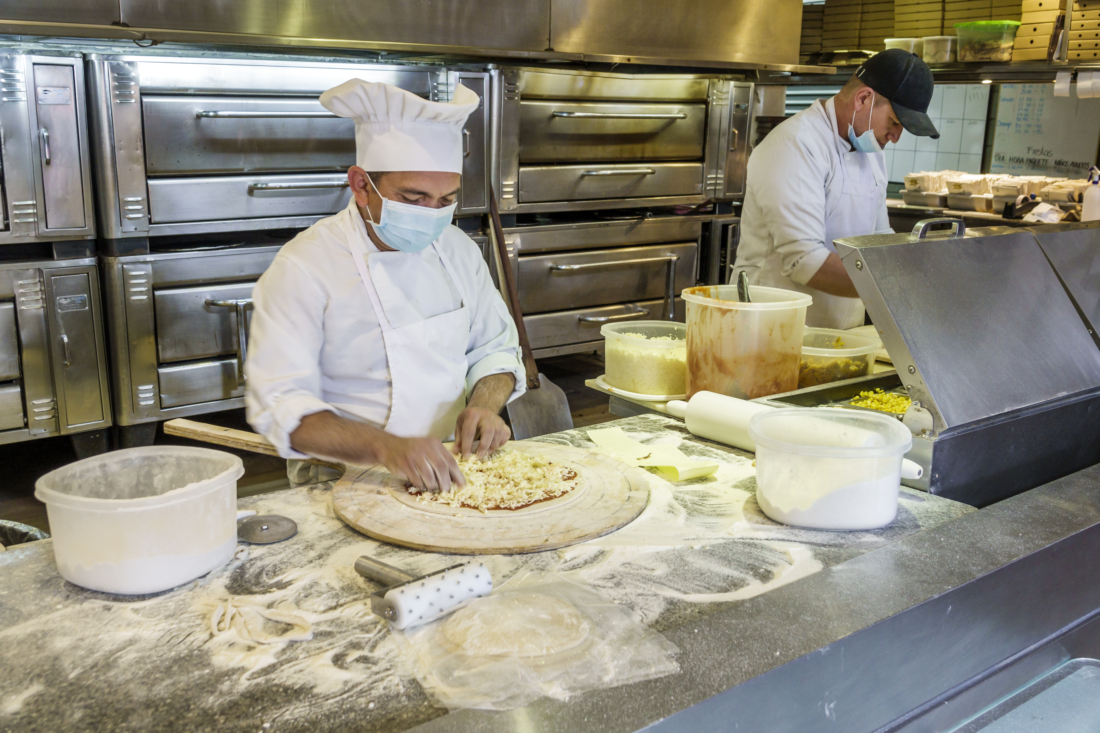 Man wearing chef’s uniform and a face mask adds topping to a pizza in a restaurant kitchen. Next to him, another man works with his mask pulled partly down his face.