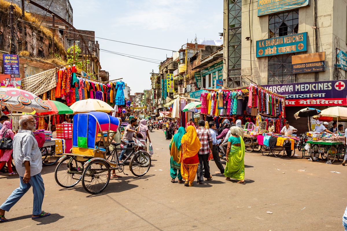 A market takes over a street in Delhi, with vendors selling colorful clothing receding into the distance in the center, an empty rickshaw to one side, and groups of locals walking.