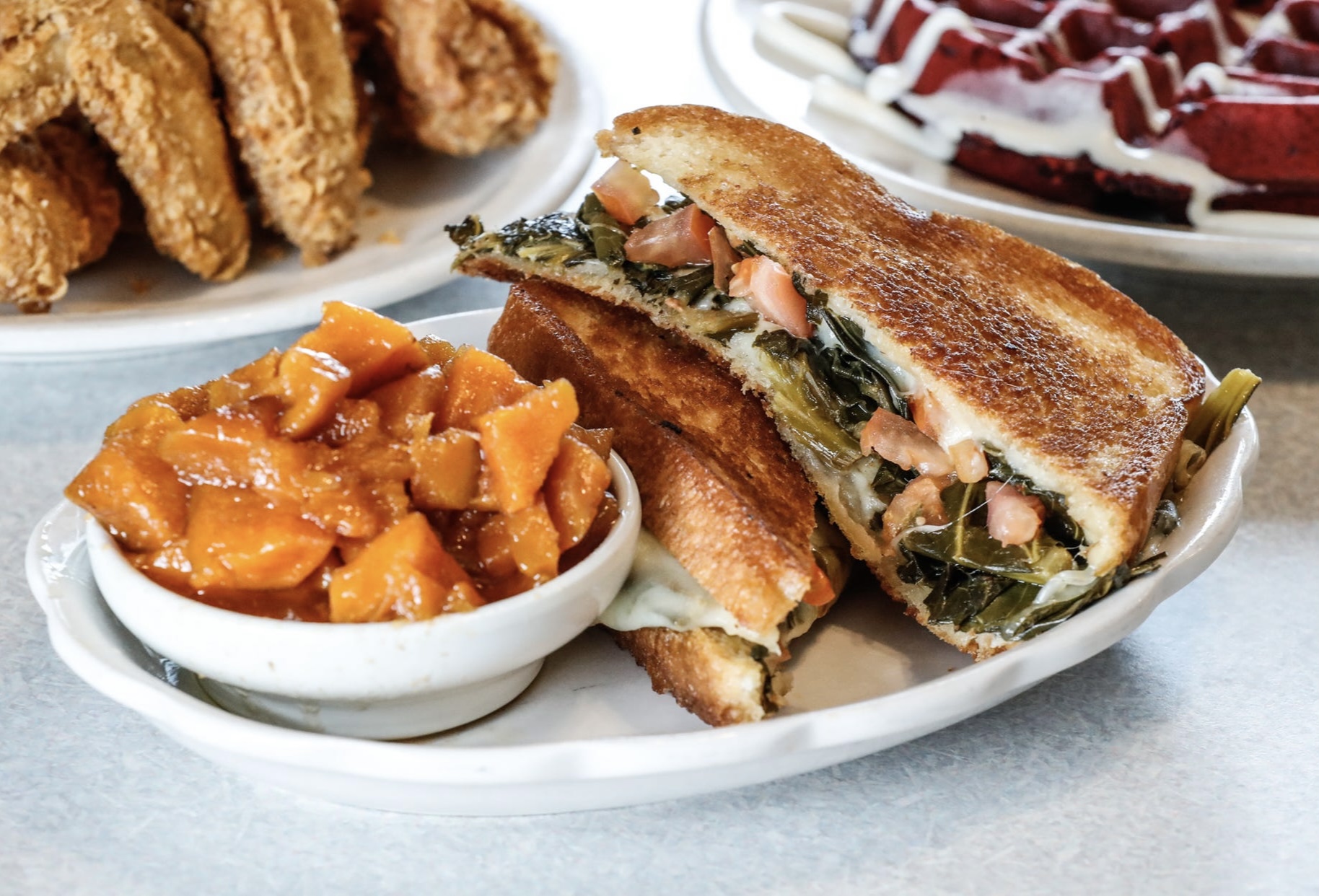 A thick grilled cheese, sliced in half and stacked on a plate beside a bowl of chunks of candied sweet potato. The sandwich bursts with collard greens, diced tomato, and cheese