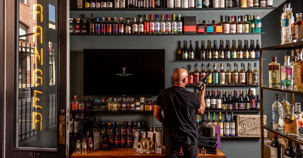 A man stands in front of a wall covered in shelves of liquor bottles and holds one in his hand. 